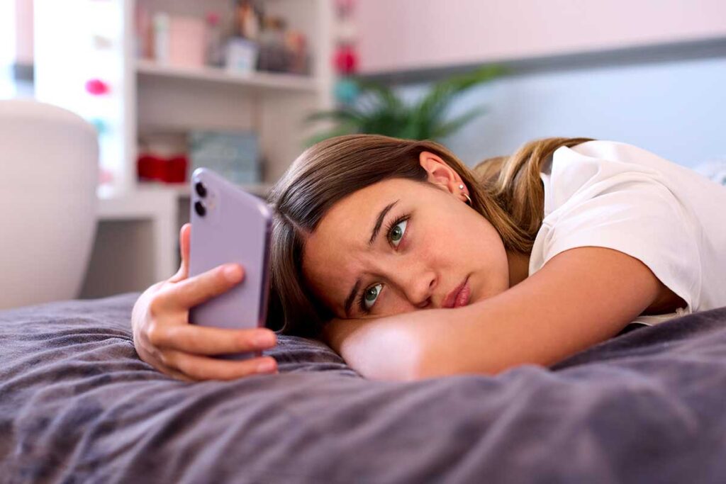 A teen girl is looking at her phone while laying in bed. Her parents wonder if this is normal teenage behavior or a mental health concern.