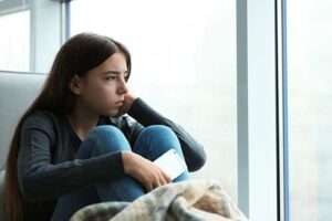An adolescent girl is staring out of her window on a dark and dreary day. She is struggling with seasonal affective disorder. 