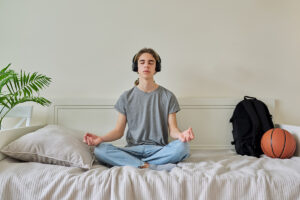 A teen is wearing headphones and practicing mindfulness and meditation while sitting cross-legged on their bed.